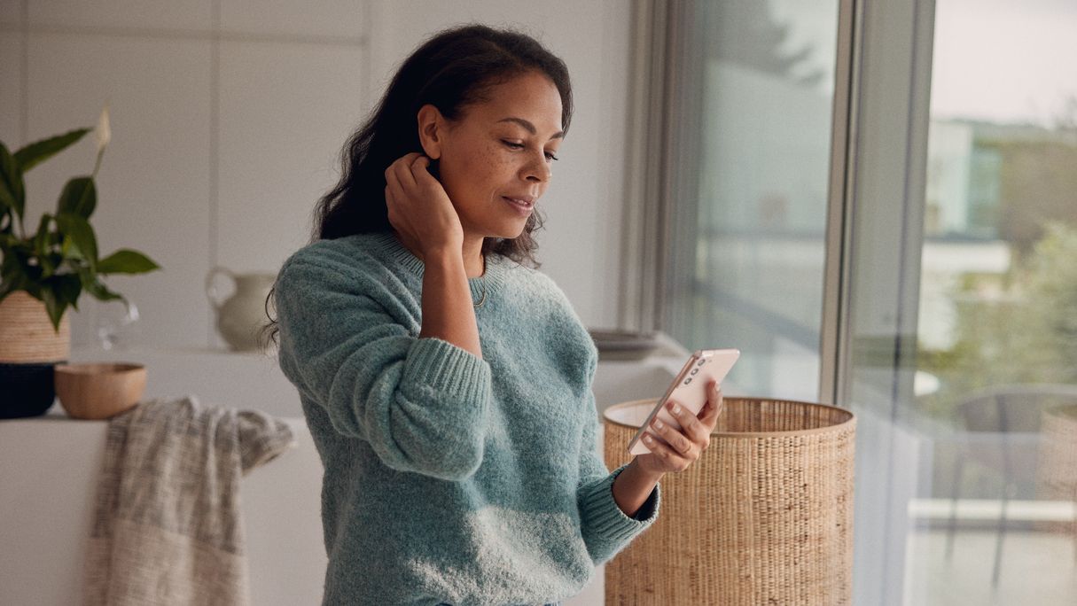 Žena stojí u okna a hledí na svůj chytrý telefon