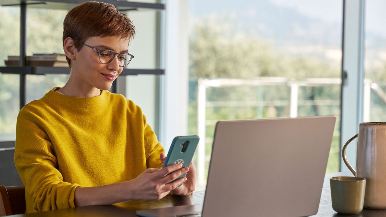 Eine Frau schaut vor ihrem Laptop auf Ihr Handy.