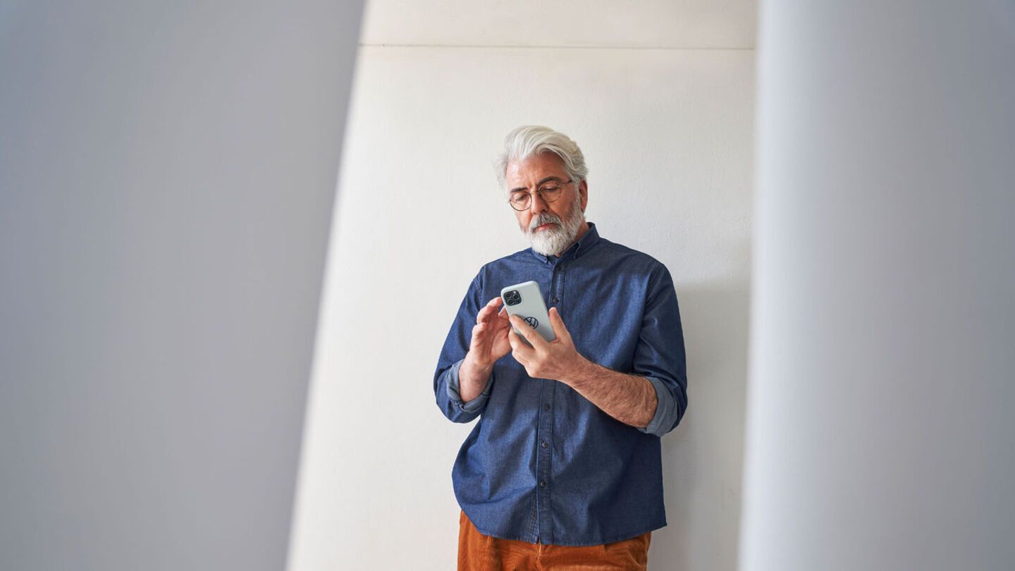 Ein Mann steht vor einer weißen Wand mit einem Smartphone in der Hand und blickt auf den Bildschirm.