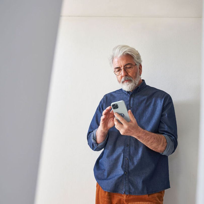 Ein Mann steht vor einer weißen Wand mit einem Smartphone in der Hand und blickt auf den Bildschirm.