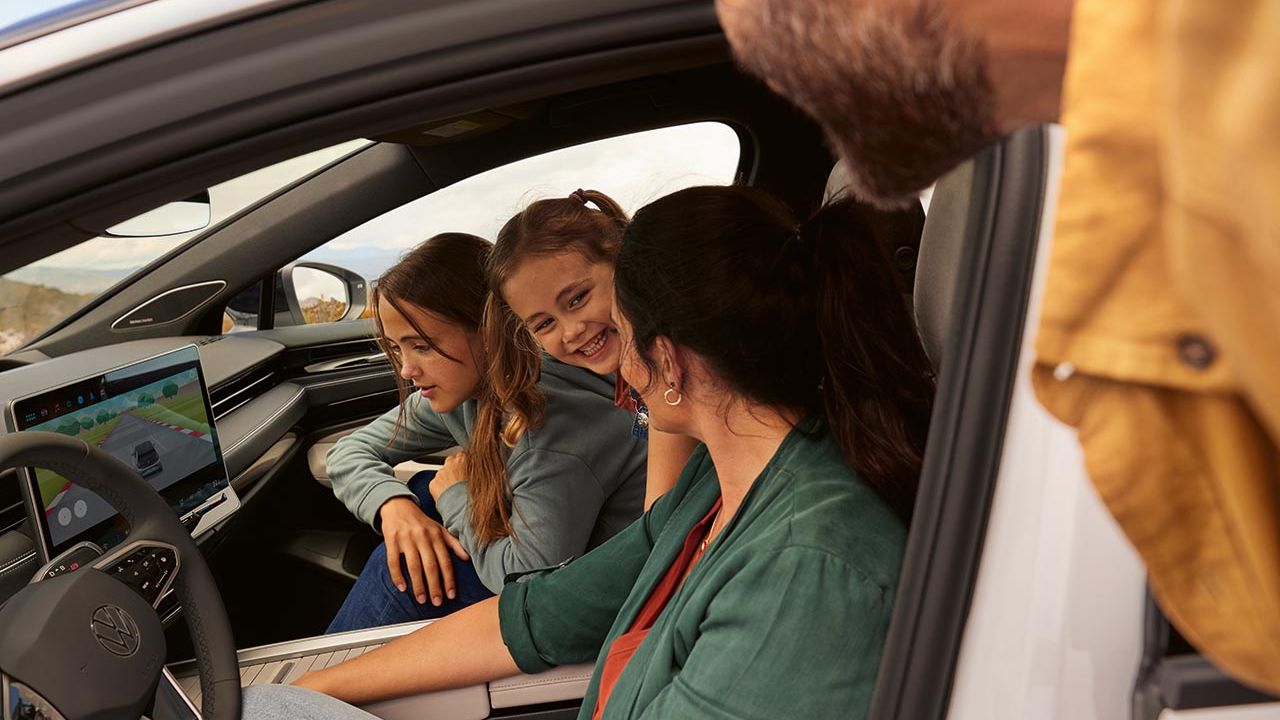 Dvě děti a žena se dívají ve stojícím voze ID.7 na systém infotainmentu, otec nahlíží dveřmi řidiče.