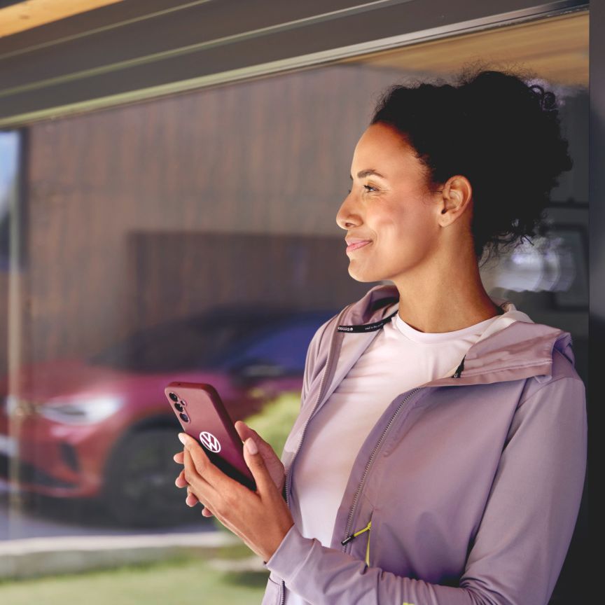 Eine Frau steht bei einem großen Glasfenster oder einer Glastür einer Hauswand. Sie hält ein Smartphone in der Hand und in der Spieglung im Glas sieht man einen roten ID.4 GTX.