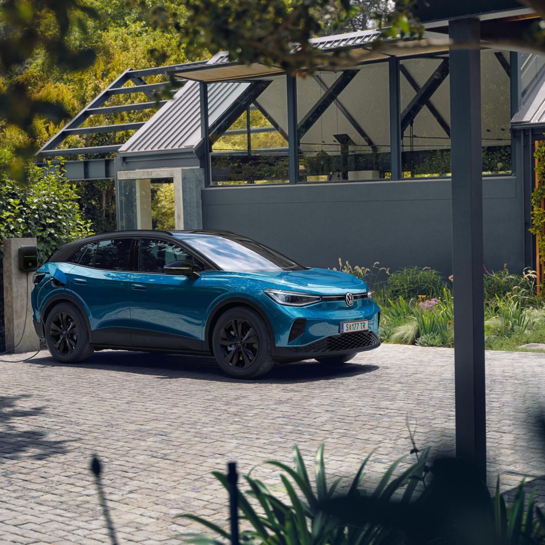 Ein blauer VW ID.4 mit schwarzem Dach parkt seitlich vor einem modernen Haus und lädt an einer Wallbox. 