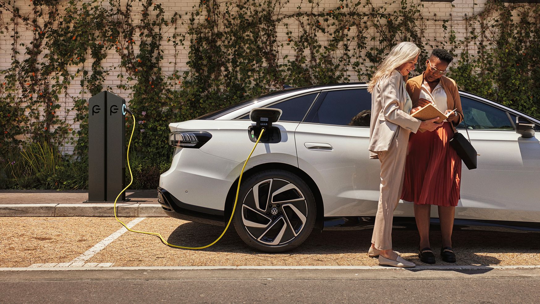 Zwei Frauen stehen mit einem Buch in der Hand neben einem weißen VW ID., der an einer öffentlichen Ladesäule lädt.