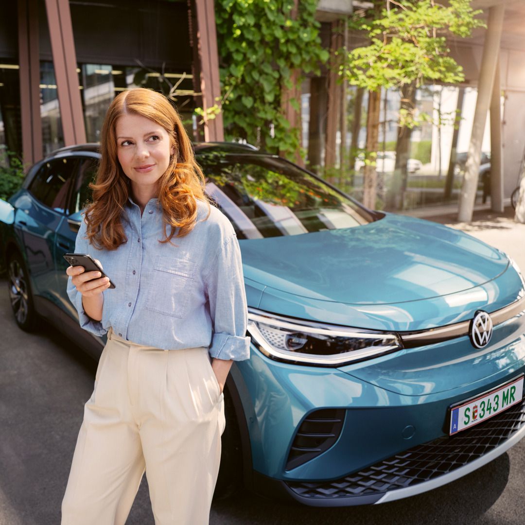 Frau mit Smartphone in der Hand steht vor der Motorhaube eines blauen VW ID.4, der an einer Wallbox angeschlossen ist.
