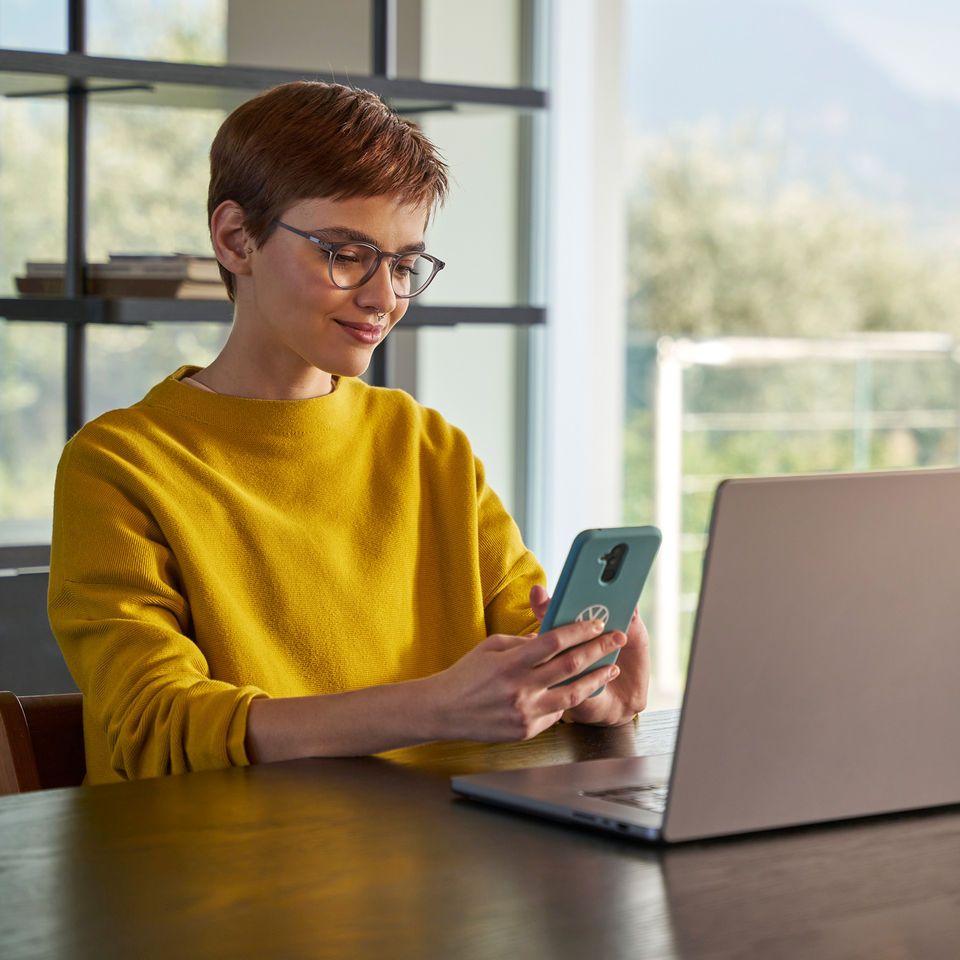 Eine Frau schaut vor ihrem Laptop auf Ihr Handy.