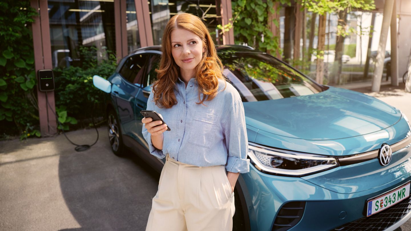 Frau mit Smartphone in der Hand steht vor der Motorhaube eines blauen VW ID.4, der an einer Wallbox angeschlossen ist.