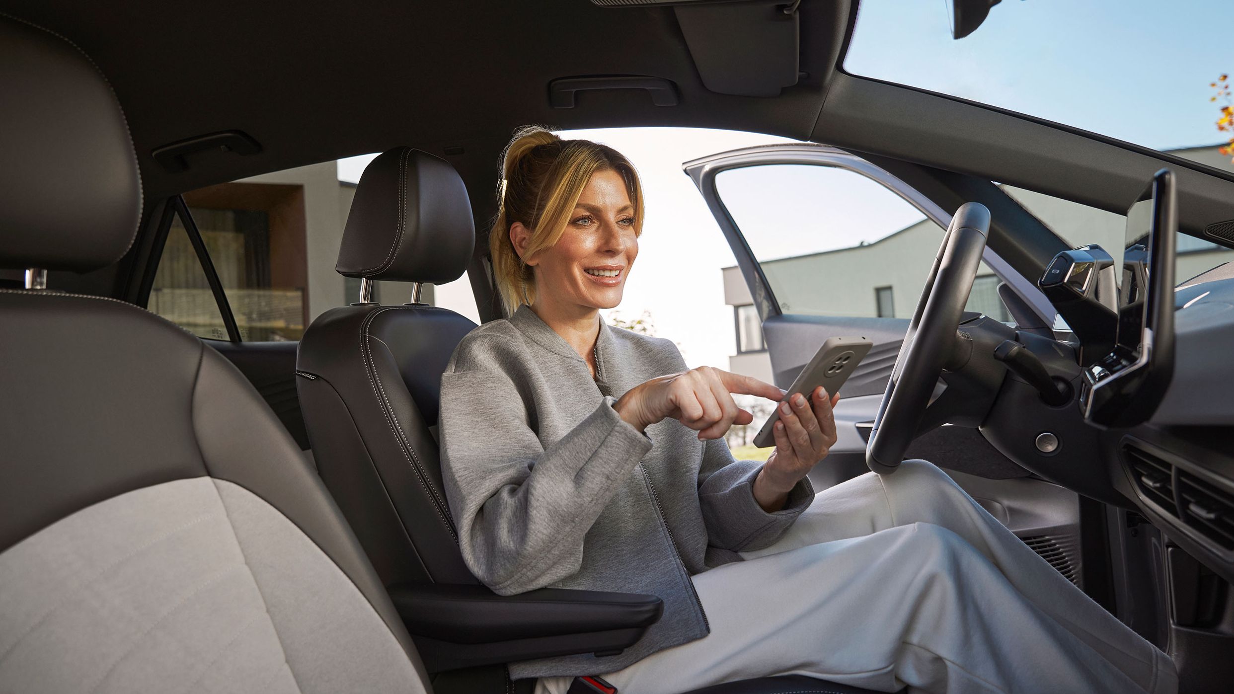 Eine Frau sitzt in einem Volkswagen auf dem Fahrersitz und bedient ein Smartphone. Der Innenraum des Autos wirkt hochwertig. 