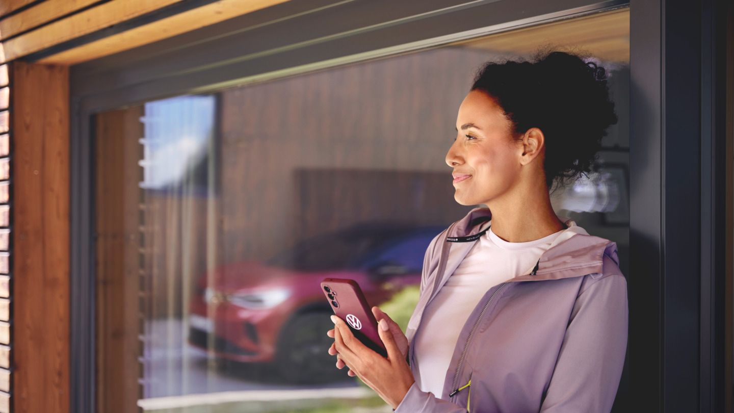 Eine Frau steht bei einem großen Glasfenster oder einer Glastür einer Hauswand. Sie hält ein Smartphone in der Hand und in der Spieglung im Glas sieht man einen roten ID.4 GTX.