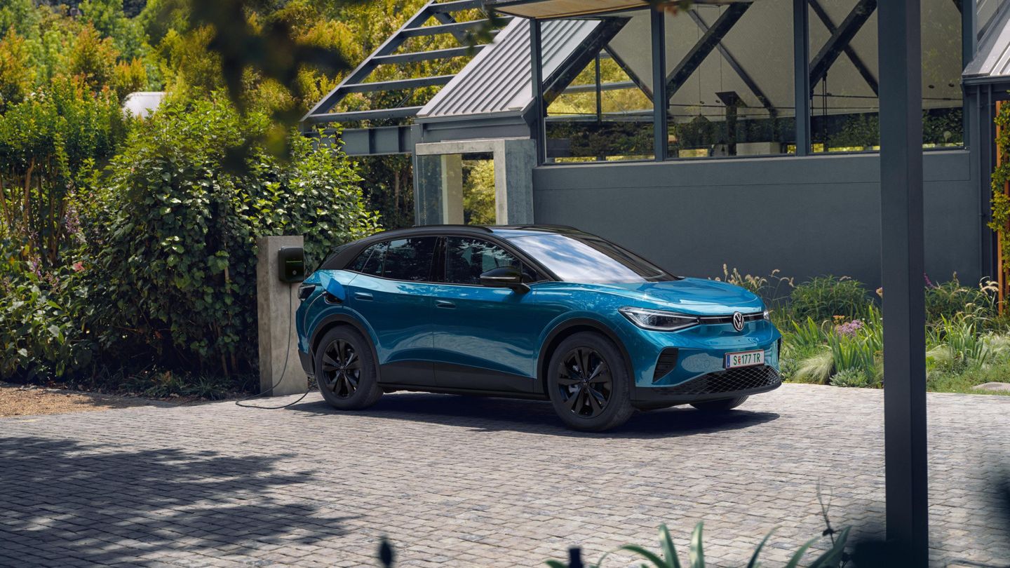Ein blauer VW ID.4 mit schwarzem Dach parkt seitlich vor einem modernen Haus und lädt an einer Wallbox. 