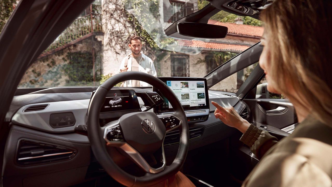 Eine Frau sitzt in einem VW und bedient den großen Touchscreen des Infotainmentsystems. Im Hintergrund steht ein Mann vor einem Haus. 