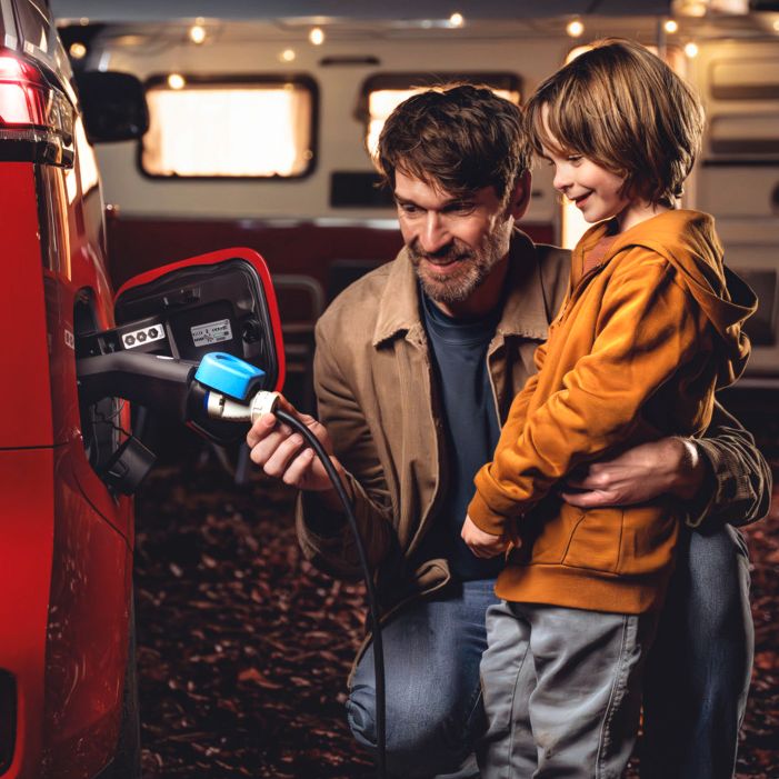Vater und Kind nutzen den ID. als Stromquelle, im Hintergrund ein beleuchtetes Wohnmobil mit Lichterkette bei Nacht.